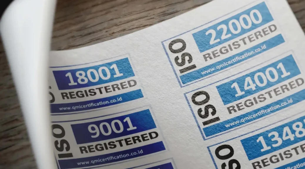 Guias de certificação com números 18001, 9001, 22000, 14001 e 13489 em selos impressos, sobre superfície de madeira, com foco em conformidade e normas industriais